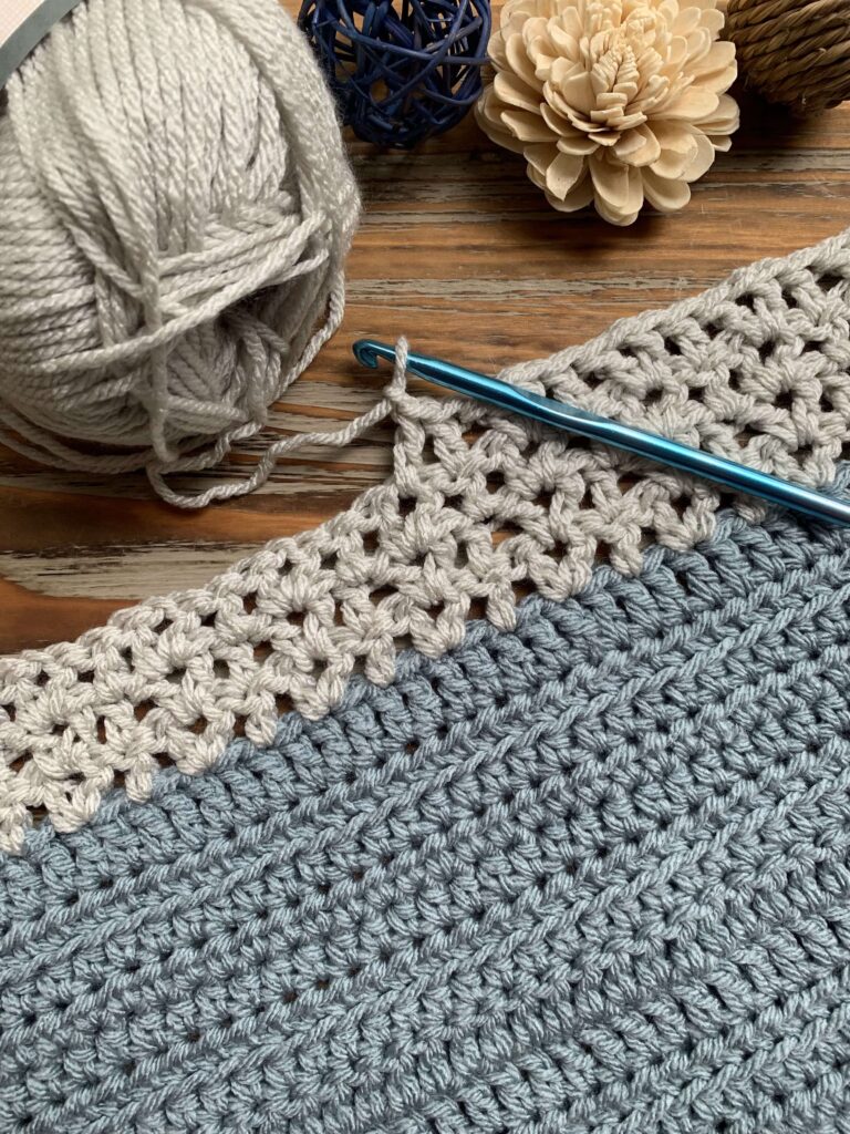 lightly textured gray and blue crochet blanket with yarn, hook, and decorative flowers